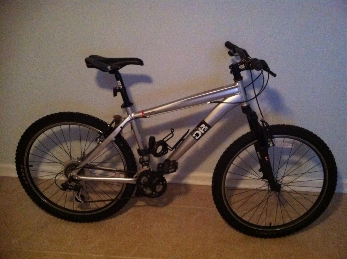 Diamondback Response: A Silver mountain bike with thick, knobby tires, set against a plain wall. The bike features a simple frame design, a black seat, and a front suspension fork, highlighting its off-road capabilities.