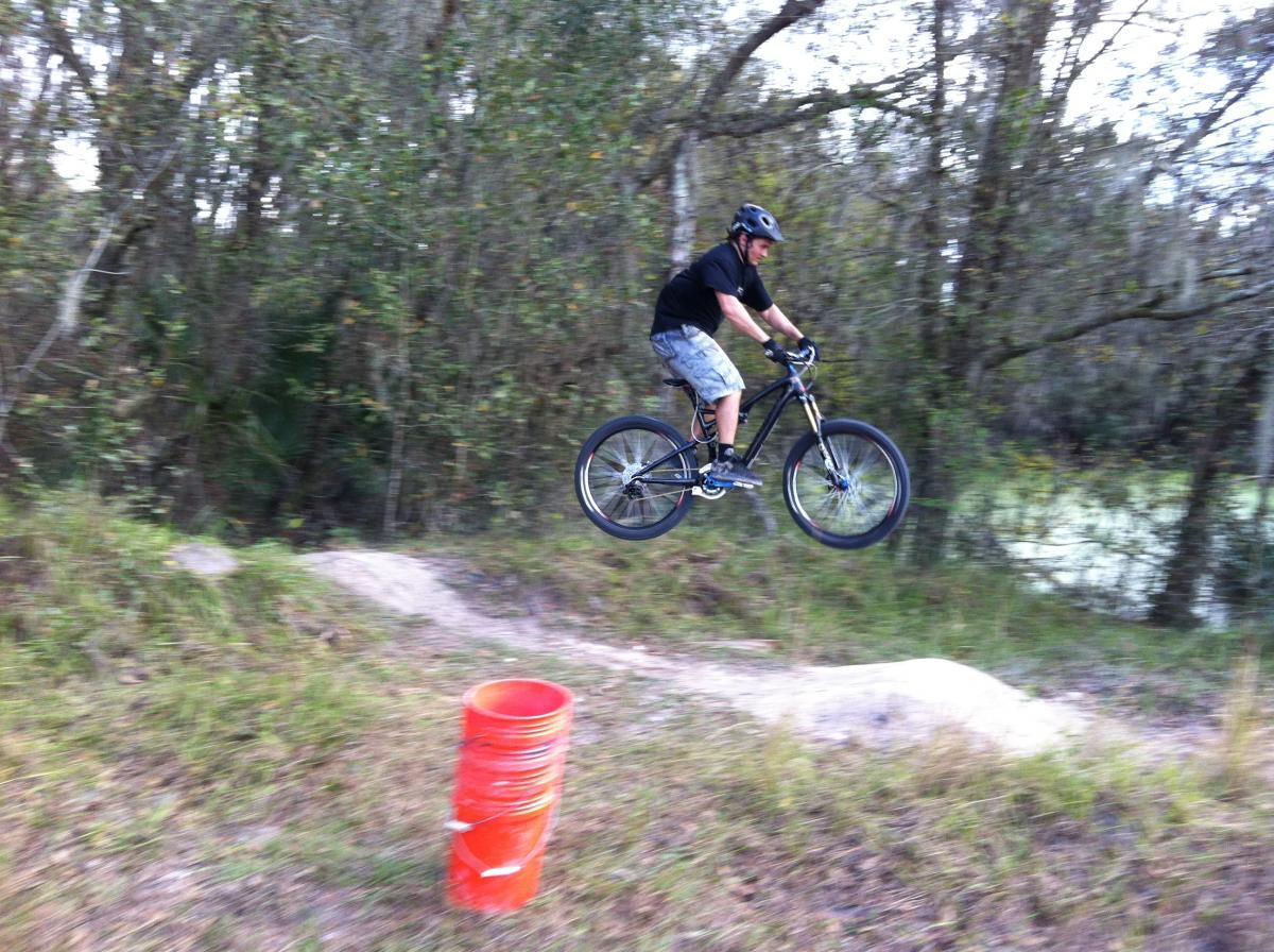 A person riding a mountain bike is airborne above a dirt ramp, surrounded by trees. An orange bucket is positioned on the ground in front of the ramp. The background features lush greenery, indicating an outdoor trail setting. Alafia River State Park mountain bike trail.