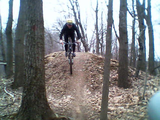 A mountain biker in a yellow helmet rides down a dirt path on a small hill, surrounded by bare trees and autumn leaves. The biker is positioned mid-air as they navigate a jump. The scene is set in a wooded area, indicating an outdoor biking trail. North Park mountain bike trail.