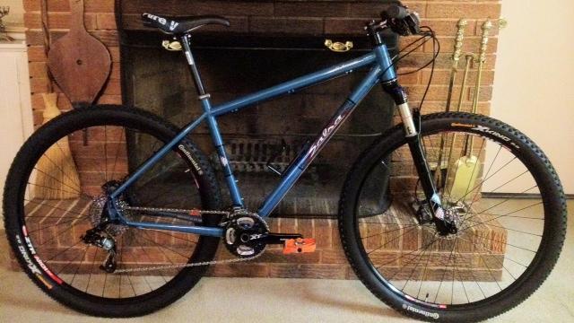 Salsa El Mariachi: A blue mountain bike with a sturdy frame and thick tires is positioned next to a brick fireplace. The bike features disc brakes and various components, including a visible chain and pedals. The background includes tools and accessories related to the fireplace, creating a warm, cozy indoor setting.