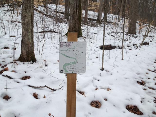 A trail map sign mounted on a wooden post in a snowy forest, with trees in the background and scattered leaves visible on the ground. The map indicates hiking routes and landmarks. Versailles State Park mountain bike trail.