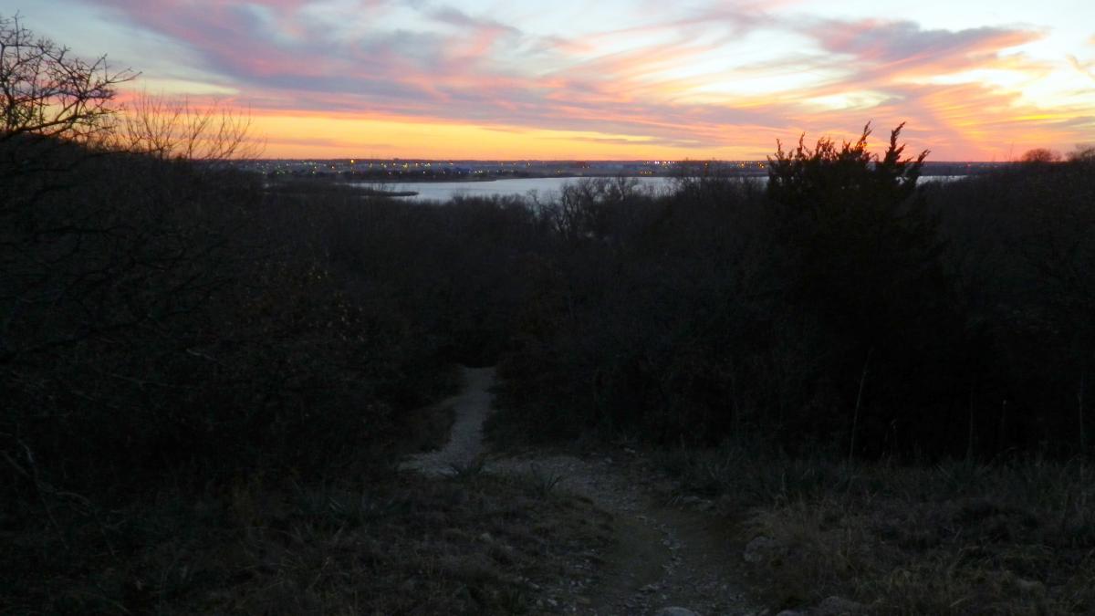 A serene twilight landscape featuring a winding dirt path leading down a slope, surrounded by trees. In the background, the sky is painted with shades of pink and orange as the sun sets, while a body of water reflects the colorful sky. The scene captures the tranquility of nature as day transitions to night. Sansom Park mountain bike trail.