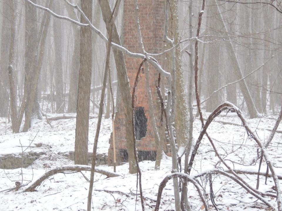 A snowy forest scene featuring a partially visible brick chimney amidst leafless trees. Light snow is falling, creating a serene, quiet atmosphere. The ground is covered in a blanket of snow, and scattered branches add to the natural, wintry landscape. Mill Stream Run Reservation mountain bike trail.