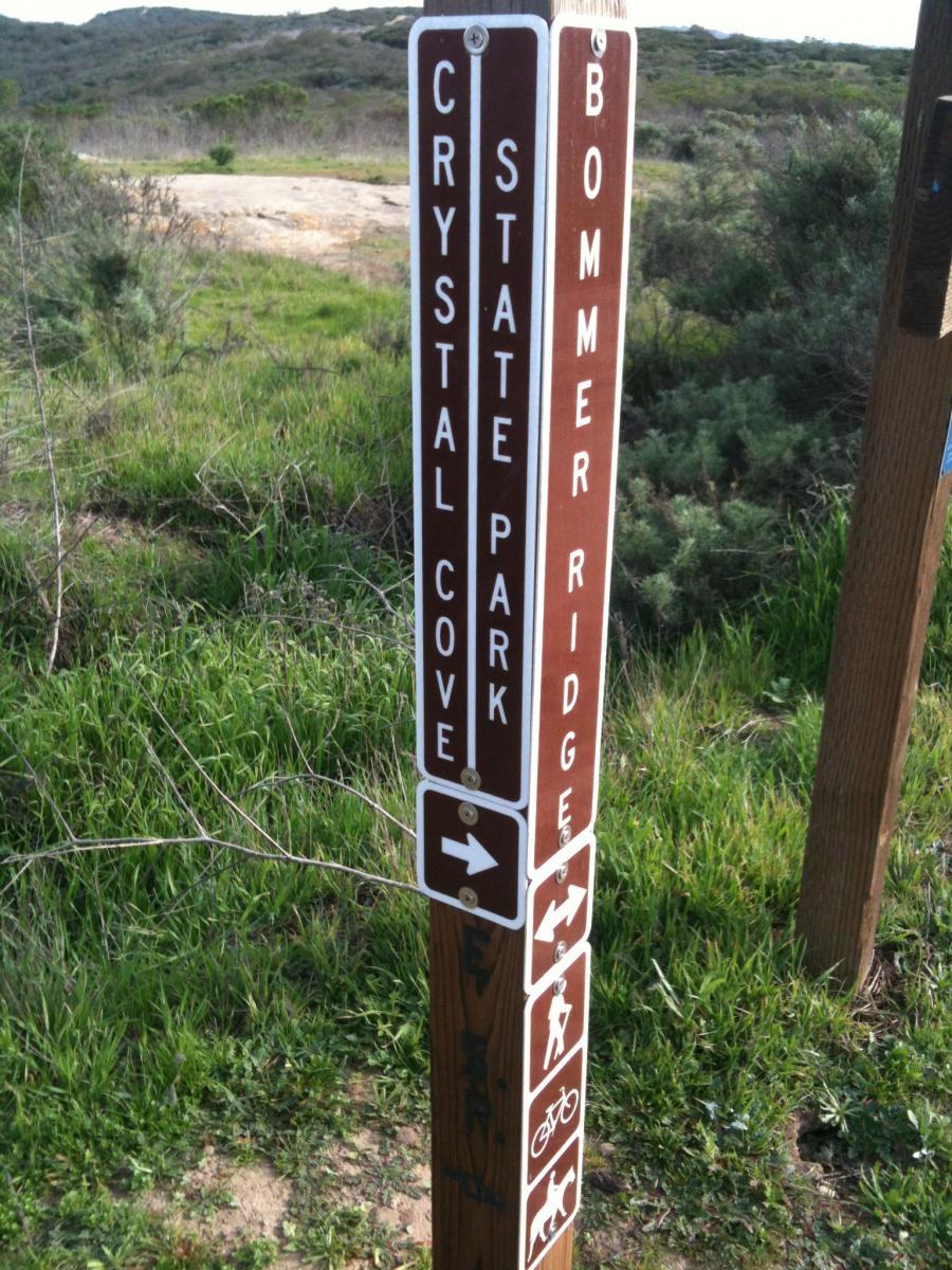 Signpost indicating trail directions and activities at Crystal Cove State Park, featuring markers for Bommer Ridge, hiking, biking, and pet-friendly paths, set against a backdrop of lush greenery and rolling hills. Crystal Cove State Park mountain bike trail.