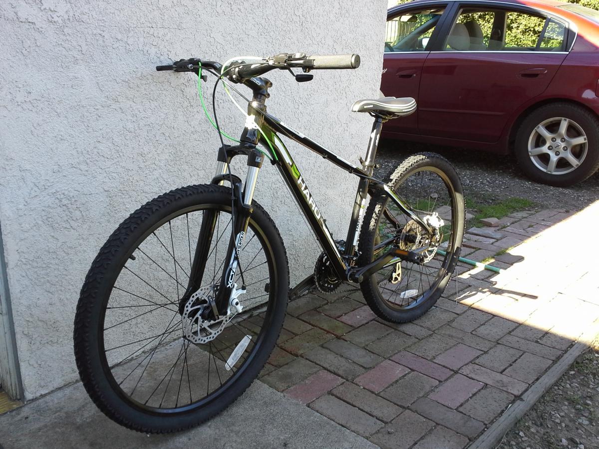 Haro Flightline Two: A black and green mountain bike leaning against a textured wall, with a red car parked in the background. The bike has thick tires and disc brakes, positioned on a paved surface with varying colors of bricks and grass peeking through.