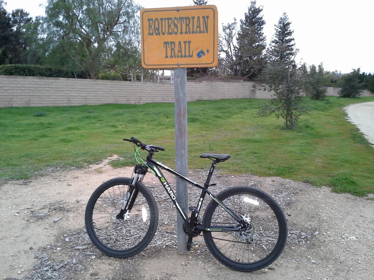 Haro Flightline Two: A mountain bike leaning against a wooden post with a sign that reads "Equestrian Trail." The background features grass, trees, and a paved path, indicating a natural outdoor setting.
