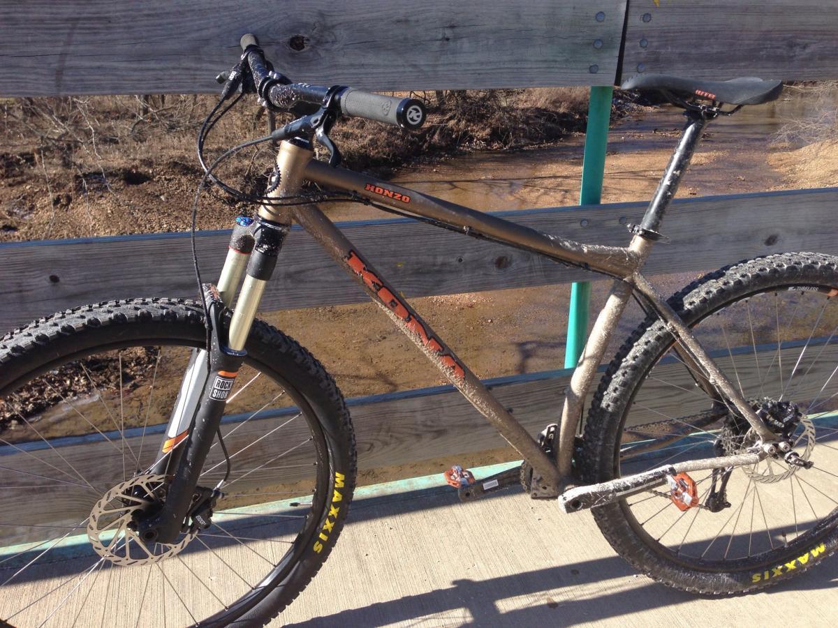 Kona Honzo: A mountain bike with a brown frame, featuring thick tires and disc brakes, parked on a wooden bridge. The background includes a view of a creek and bare trees, indicating an outdoor setting. The bike appears to have some dirt splatter, suggesting recent use on a trail.