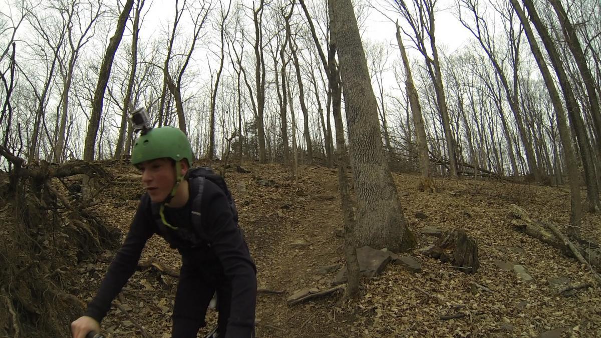 A person wearing a green helmet rides a mountain bike on a wooded trail. The surroundings feature bare trees and dried leaves on the ground, indicating an early spring or late autumn setting. High Rocks mountain bike trail.