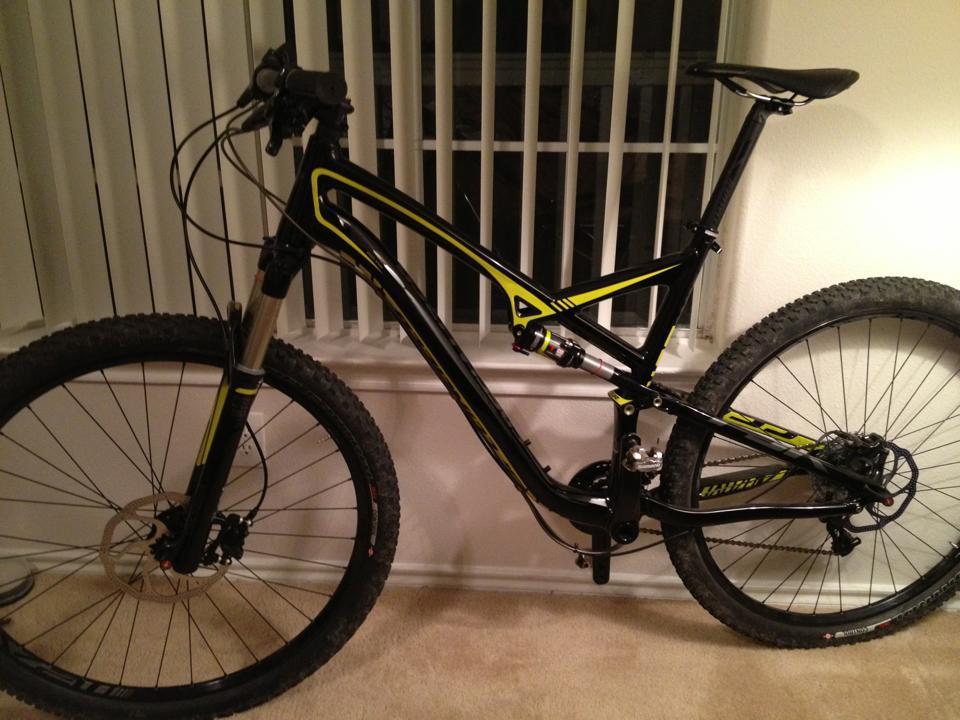 Specialized Camber: A black and yellow mountain bike stands upright against a wall, positioned on a carpeted floor. The bike features thick tires and a suspension system, with its handlebars turned slightly to the side. In the background, there are vertical blinds allowing some light to filter through.