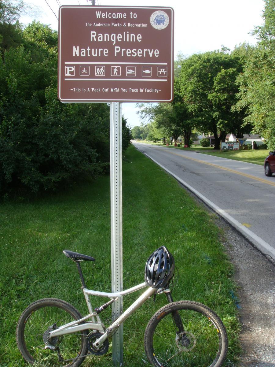 A welcome sign for Rangeline Nature Preserve, featuring symbols for parking, biking, hiking, and picnicking. A silver mountain bike with a black helmet rests against the sign, set against a backdrop of green grass and trees along a road. Rangeline Nature Preserve mountain bike trail.