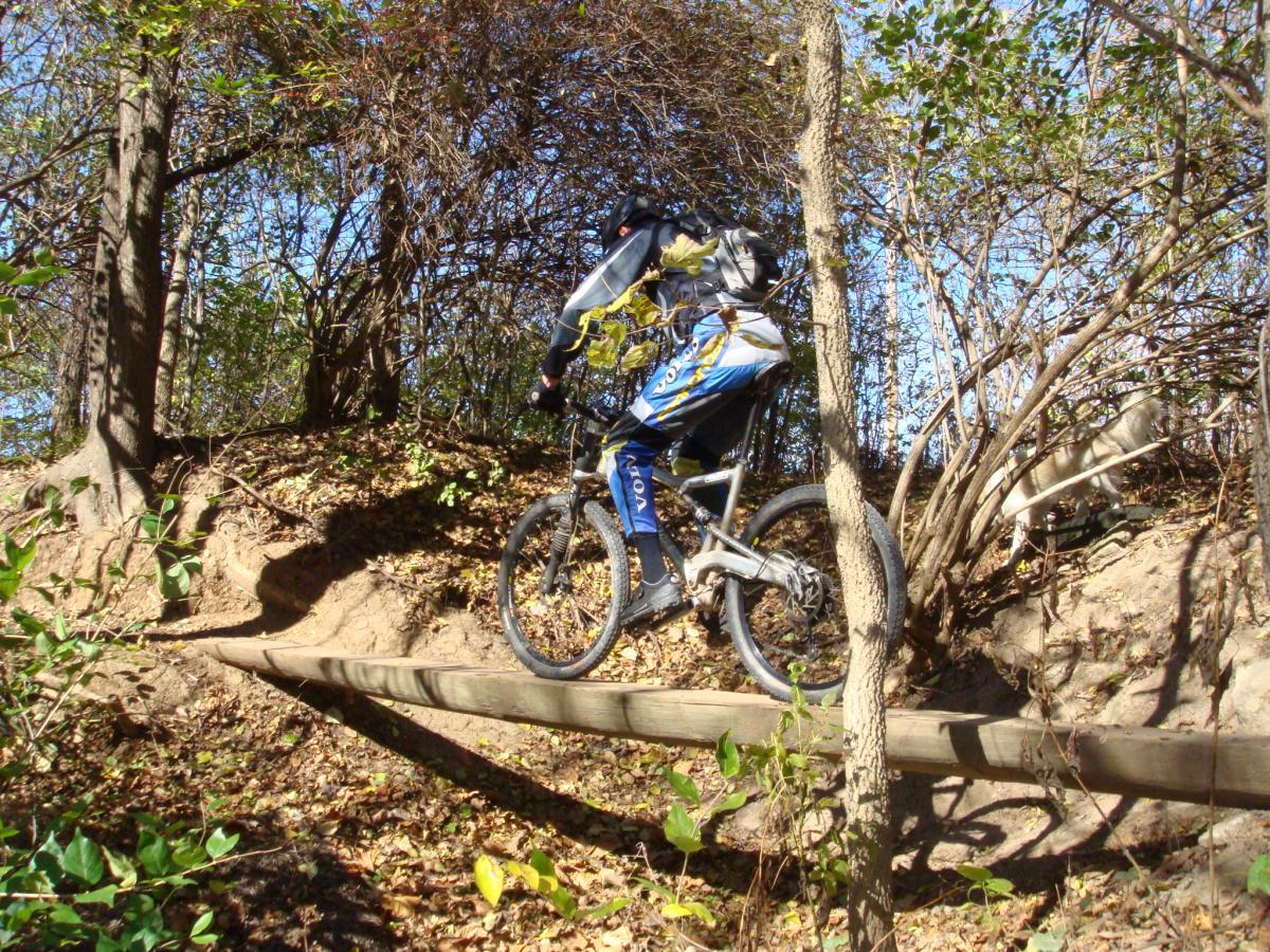 A mountain biker navigating a narrow wooden bridge through a wooded area, surrounded by trees and autumn foliage. Rangeline Nature Preserve mountain bike trail.