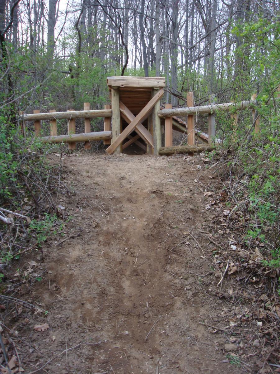 A dirt path leading to a wooden trail structure surrounded by trees and greenery, featuring a rustic wooden barrier with an X brace. Rangeline Nature Preserve mountain bike trail.