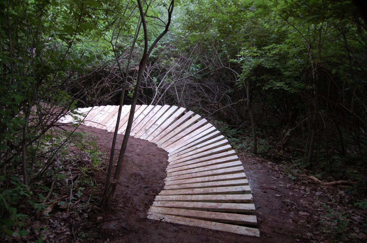 A curving wooden path made of slats, set in a forested area with dense greenery and underbrush surrounding it. The path is partially covered with dirt and leads deeper into the woods. Rangeline Nature Preserve mountain bike trail.