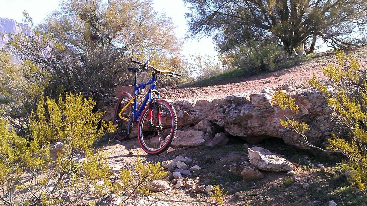 A blue and yellow mountain bike is leaning against a rocky outcrop in a desert landscape, surrounded by sparse greenery and shrubs. The scene is sunlit, showcasing a trail winding through the rough terrain. Cloudview mountain bike trail.