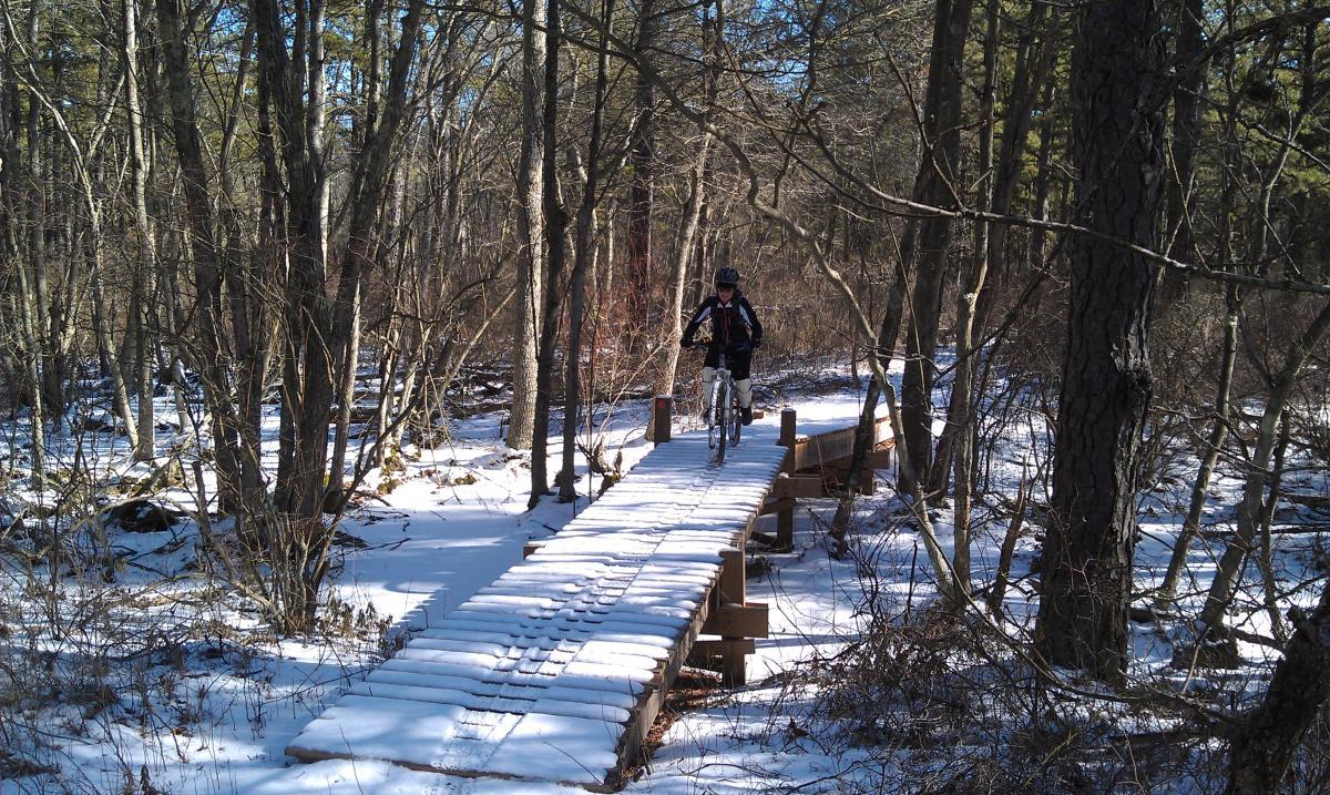 A cyclist rides a mountain bike across a wooden bridge surrounded by snow-covered ground and trees in a winter forest setting. Wharton State Forest mountain bike trail.