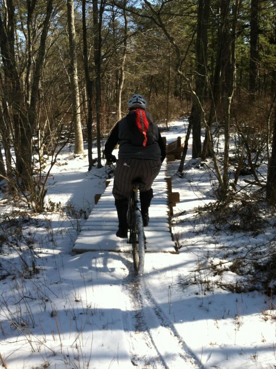 A person biking over a wooden bridge in a snowy forested area, surrounded by trees and a clear blue sky. The cyclist is wearing a helmet and has a red scarf, with tire tracks visible in the snow along the path. Wharton State Forest mountain bike trail.