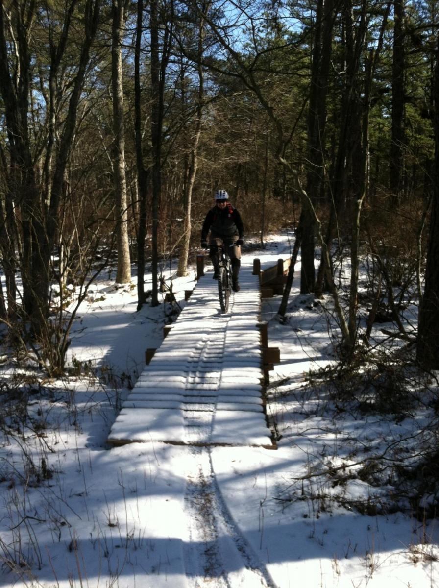 A person riding a mountain bike over a wooden plank bridge in a snowy, wooded area. Sunlight filters through the trees, casting shadows on the snow-covered ground and the bridge. Wharton State Forest mountain bike trail.