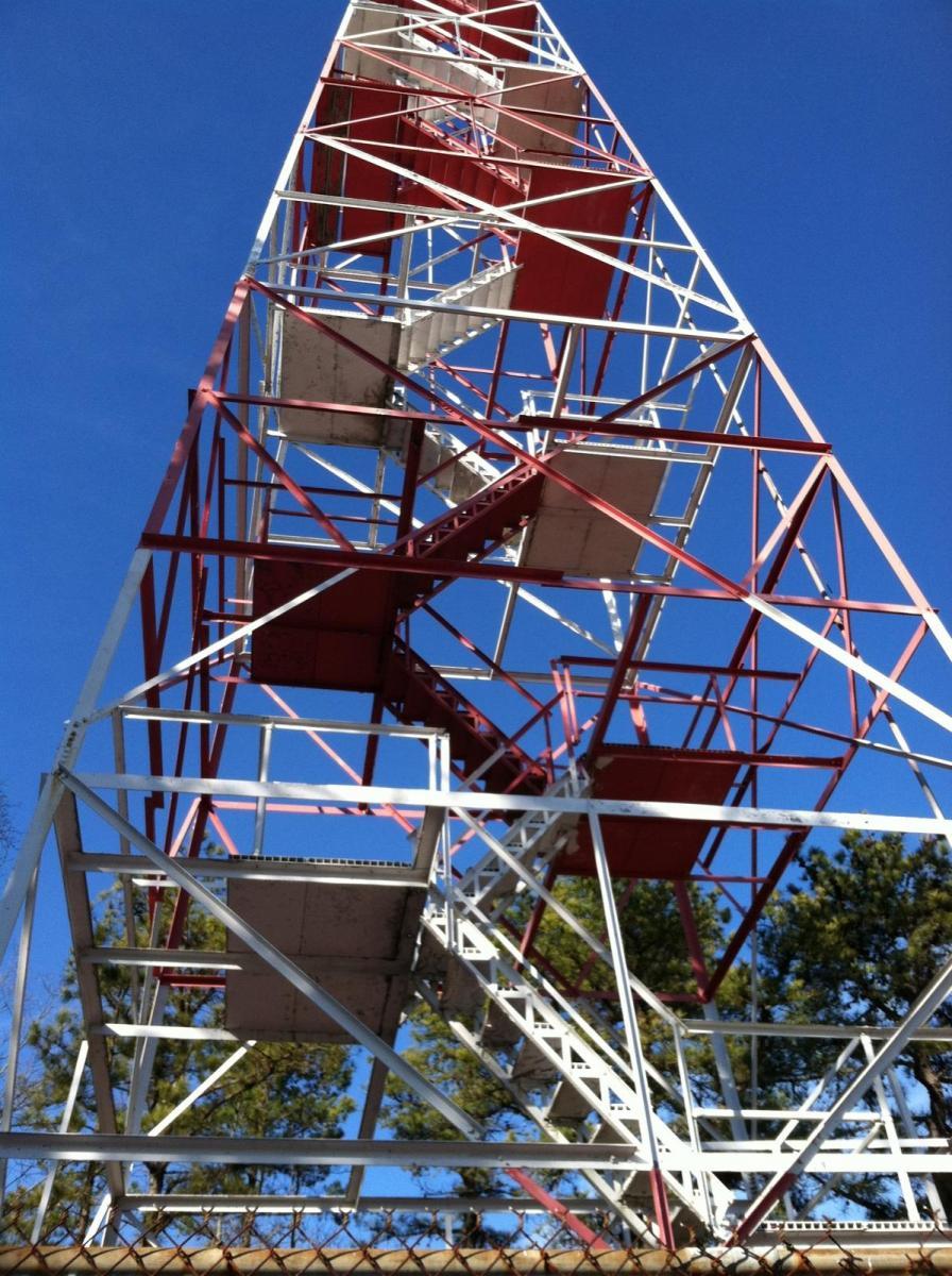 A red and white metal tower structure viewed from below against a clear blue sky, with visible steps and platforms, surrounded by trees and a chain-link fence at the base. Wharton State Forest mountain bike trail.