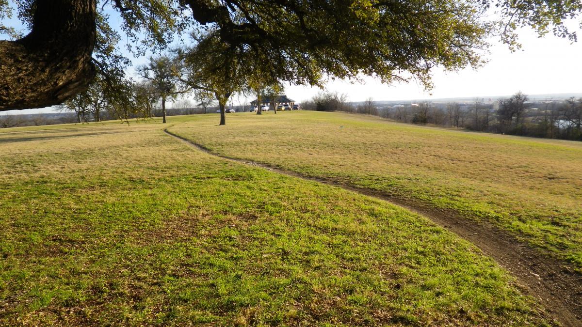 A scenic view of a grassy hill under a clear sky, featuring a winding dirt path leading toward a structure in the distance, surrounded by trees. Sansom Park mountain bike trail.