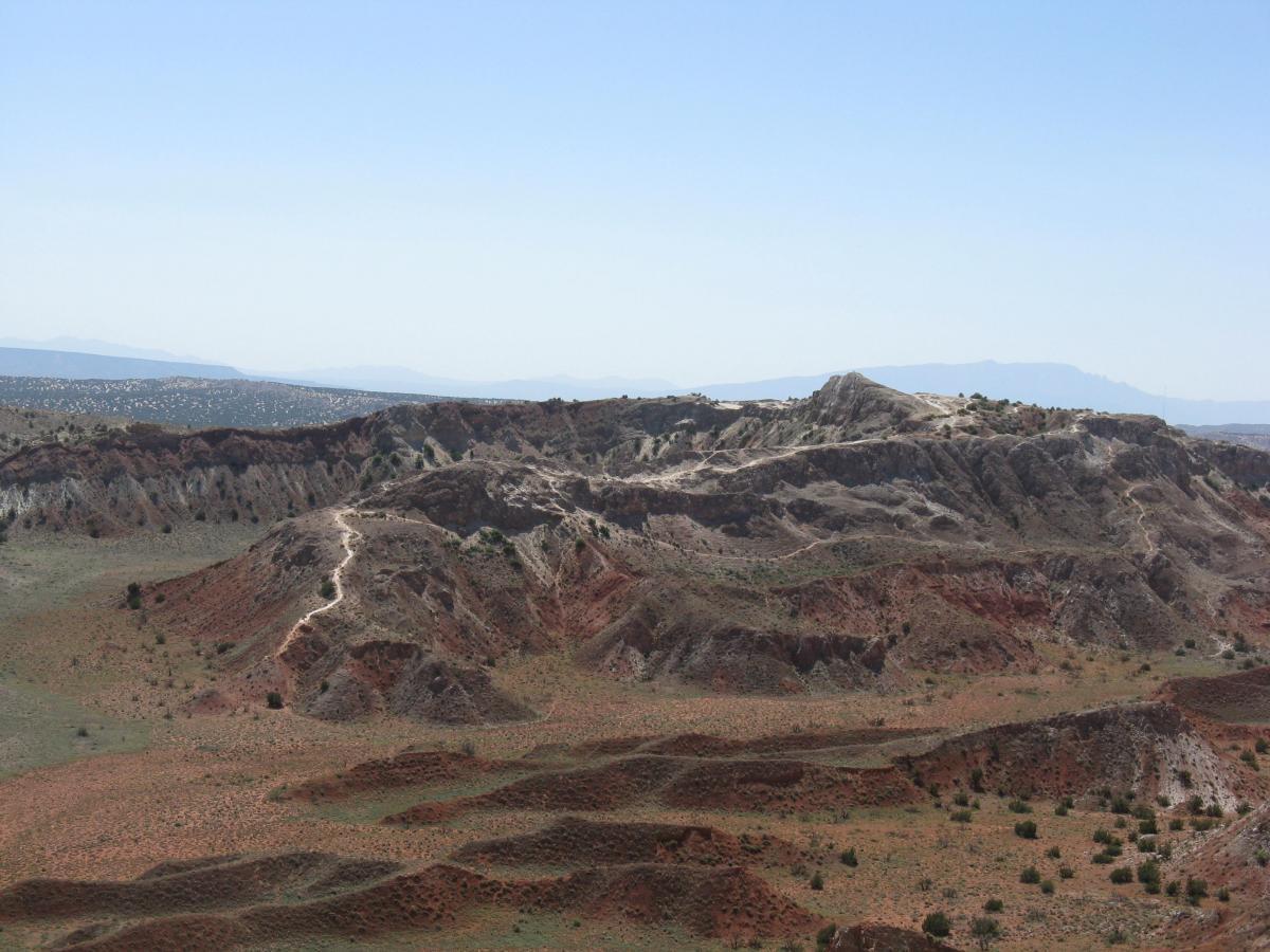 A panoramic view of rugged hills and textured land formations, featuring layers of red and gray earth under a clear blue sky. The landscape includes visible hiking trails winding through the terrain, with distant mountains on the horizon. White Ridge Bike Trails mountain bike trail.