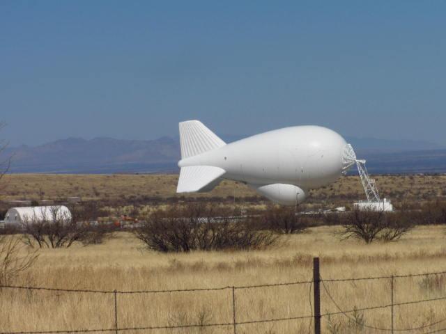 A large, white blimp-like structure is situated in a grassy field, with mountains visible in the background. The structure has a streamlined shape and features tail fins. A small building is visible nearby, along with some sparse vegetation and a barbed-wire fence in the foreground. The scene is set under a clear blue sky. Brown Canyon mountain bike trail.