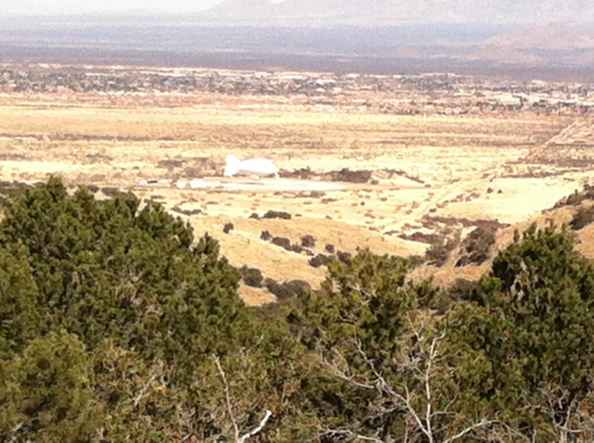 A landscape view from a hillside overlooking a vast, arid valley with sparse vegetation and distant mountains. In the foreground, green shrubs and trees frame the scene, while a white structure is faintly visible in the middle ground against the dry terrain. The sky is clear with a hint of haze in the distance. Brown Canyon mountain bike trail.