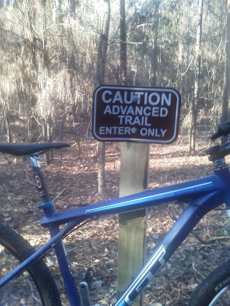 A blue mountain bike is leaning against a wooden sign in a wooded area. The sign reads "CAUTION ADVANCED TRAIL ENTER ONLY." The ground is covered with fallen leaves, and trees are visible in the background, creating a natural trail setting. Tillie Fowler Regional Park mountain bike trail.