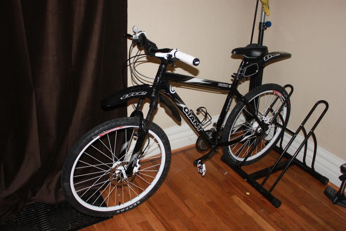 Giant Yukon: A black mountain bike with white accents, mounted on a bike stand, positioned beside a brown curtain in a room with hardwood flooring.