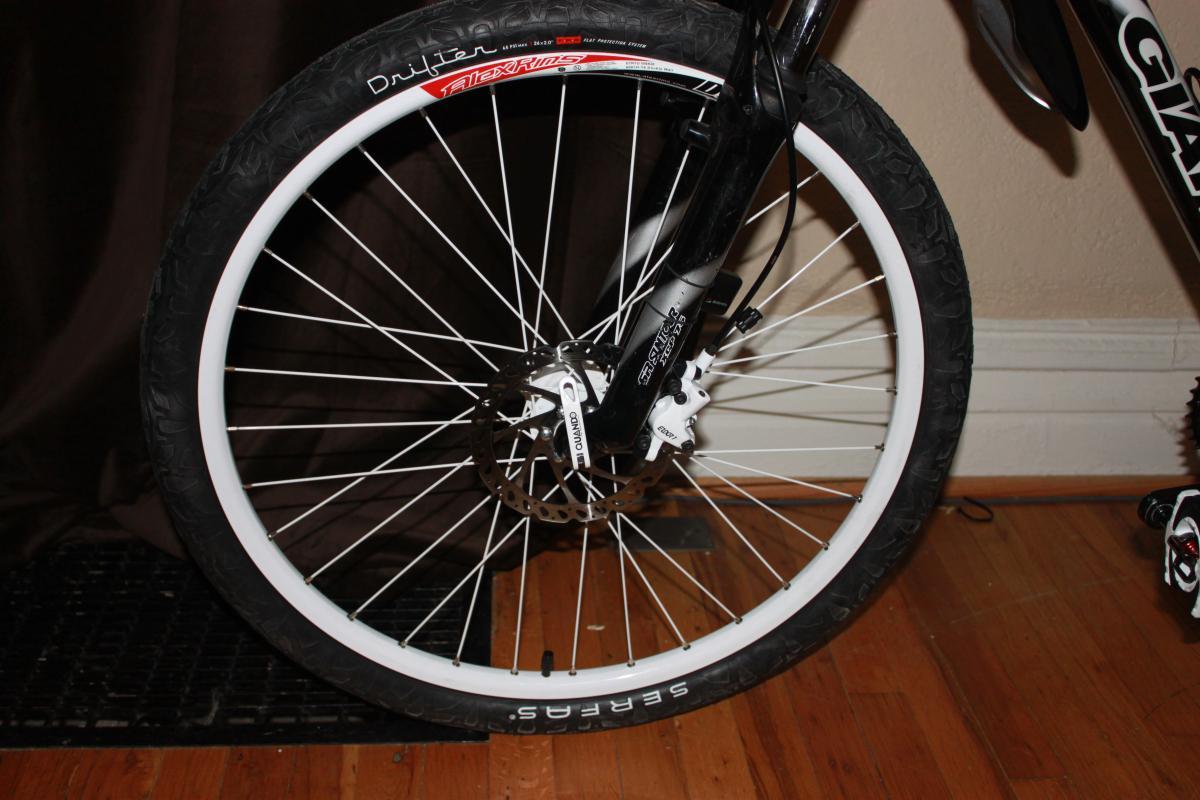 Giant Yukon: Close-up image of a bicycle front wheel with a white rim and a black tire, showcasing the spokes and brake disc. The background features wooden flooring and a dark curtain.