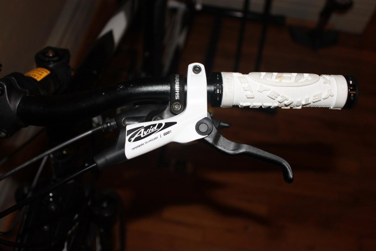 Giant Yukon: Close-up of a bike handlebar featuring a white Avid brake lever and a textured white handlebar grip. The brake lever is positioned near the frame, and there are visible cables and components associated with the braking system. The background shows wooden flooring and part of the bike frame.