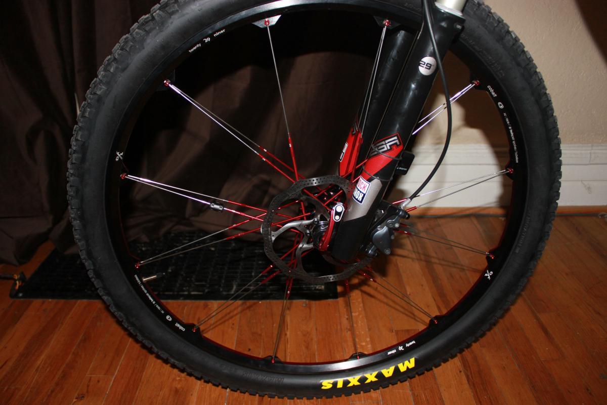 Scott Spark 29 Elite: Close-up view of a bicycle wheel featuring a black tire with a prominent "Maxxis" label, red spokes, and a disc brake system. The background includes a dark curtain and wooden flooring.