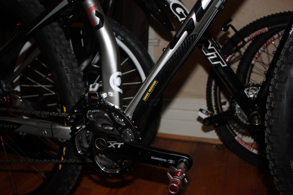 Scott Spark 29 Elite: Close-up of a bicycle's drivetrain and frame, featuring Shimano XT components and a carbon fiber section, with visible branding and multiple bicycles in the background. The floor is wooden, and the setting appears to be an indoor space.