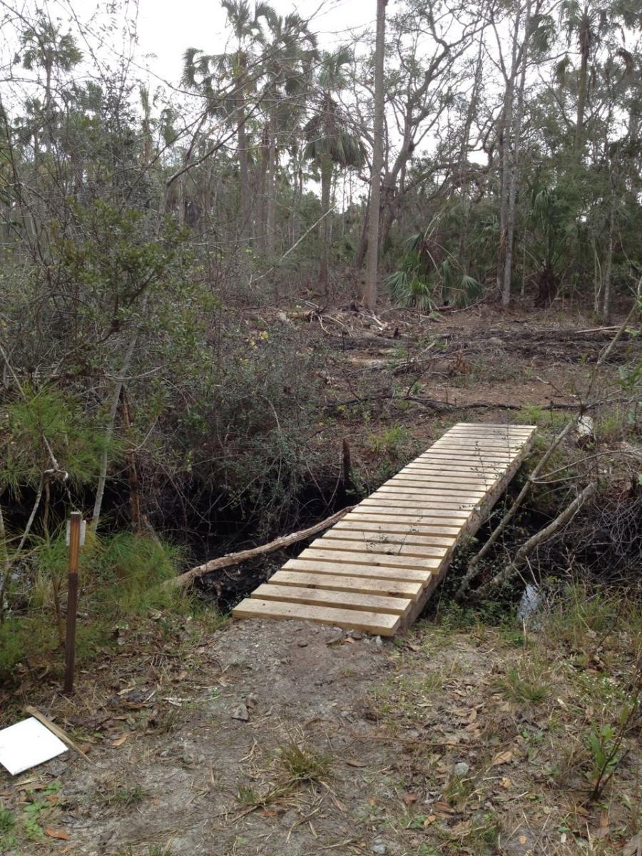 A wooden boardwalk spans a narrow gap in a forested area, surrounded by dense underbrush and trees, with some recent clearing visible in the background. A post with a marker stands to the left, and a white sign lies on the ground nearby. The scene is bright but overcast, indicating a natural environment still in the process of rehabilitation. Nocatee mountain bike trail.