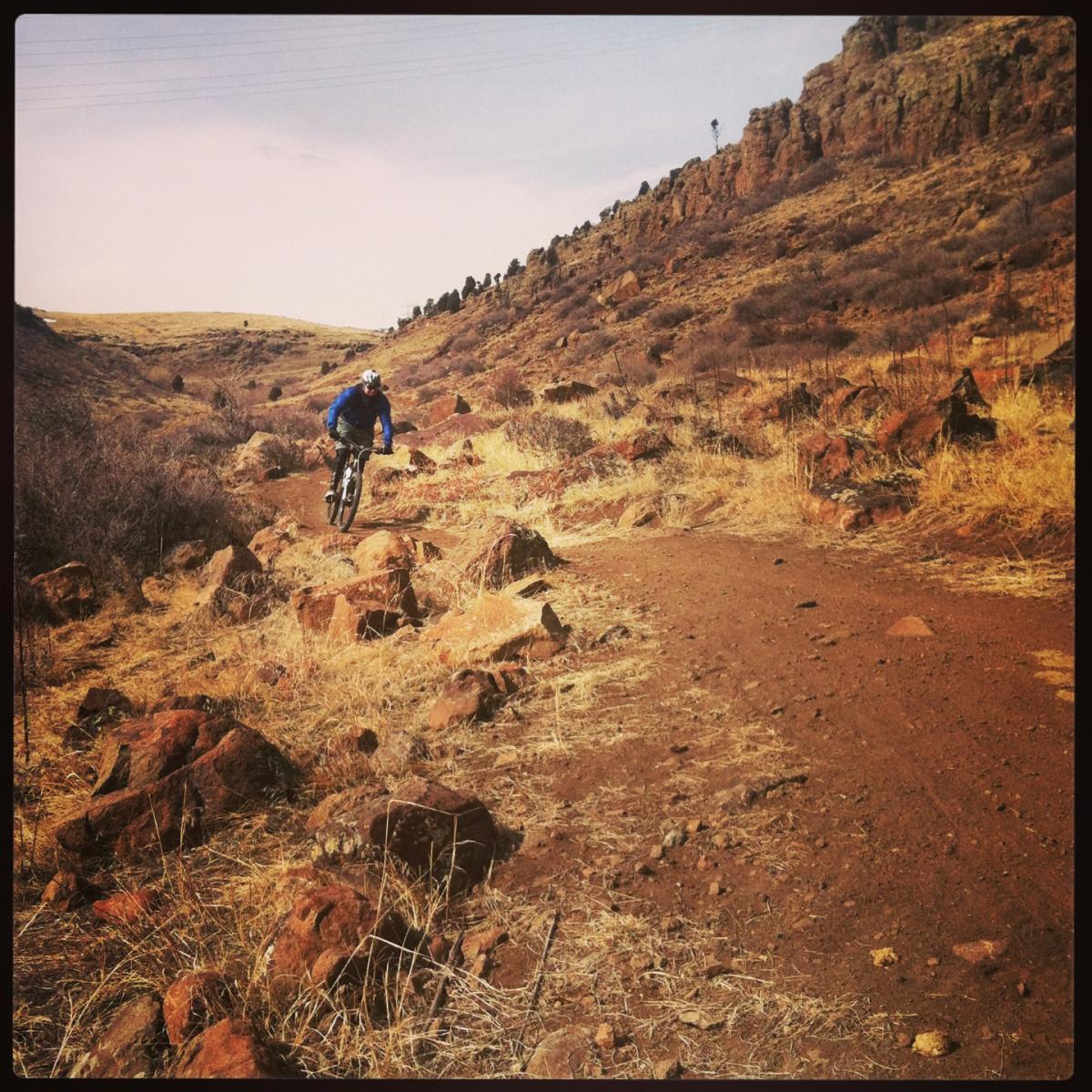A mountain biker rides along a rugged path surrounded by rocky terrain and dry grass. In the background, rolling hills can be seen under a clear sky. North Table Mountain mountain bike trail.