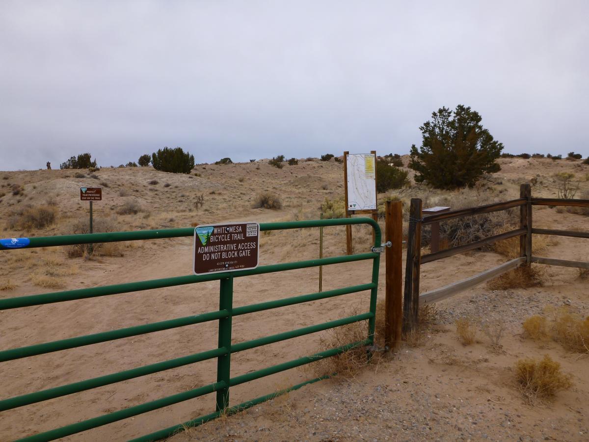 A green gate at the entrance to the Whit Mesa Bicycle Trail, surrounded by sandy terrain and sparse vegetation. A sign on the gate indicates "Administrative Access - Do Not Block Gate," and there is a trail map mounted on a nearby post. In the background, a desolate landscape extends under a cloudy sky. White Ridge Bike Trails mountain bike trail.