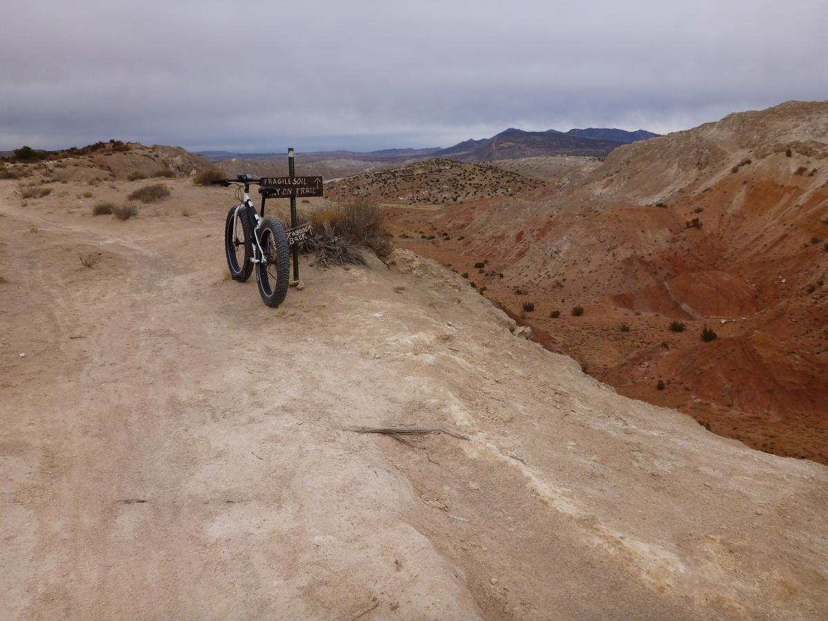 A mountain bike rests on a trail overlooking a rugged landscape with colorful rock formations and sagebrush. A sign labeled "Fragile Soil" points towards the trail ahead, under a cloudy sky. The scene captures a remote, natural environment, ideal for outdoor adventures. White Ridge Bike Trails mountain bike trail.