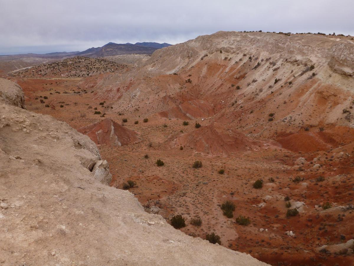 A panoramic view of a rugged, dry landscape featuring reddish-brown hills and valleys under a cloudy sky. Sparse vegetation is visible among the arid terrain, with distant mountains lining the horizon. The scene captures the unique geological formations of the area. White Ridge Bike Trails mountain bike trail.