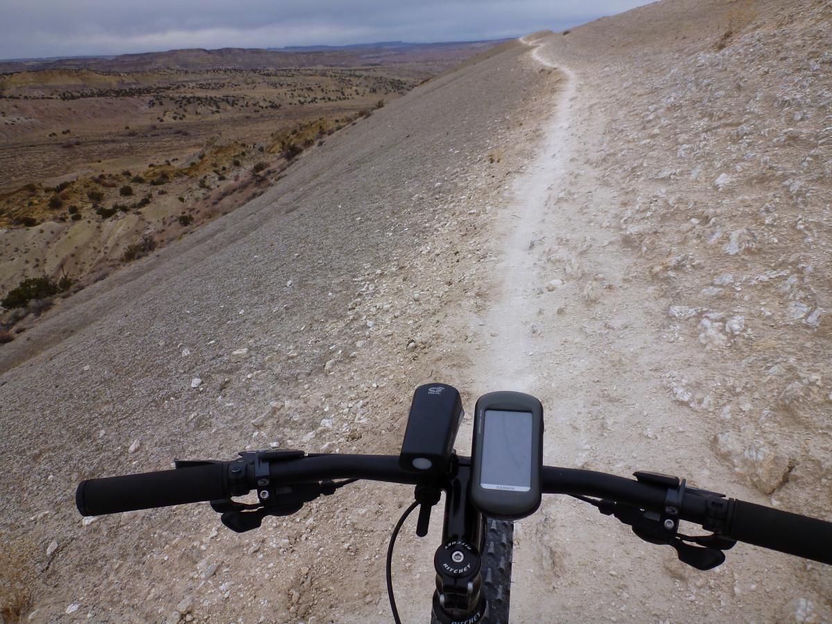 A view from the handlebars of a mountain bike on a narrow, rocky trail. The path winds along a rugged landscape with hills and valleys in the background, under a cloudy sky. A bicycle computer is visible on the handlebars, displaying speed and distance metrics. White Ridge Bike Trails mountain bike trail.