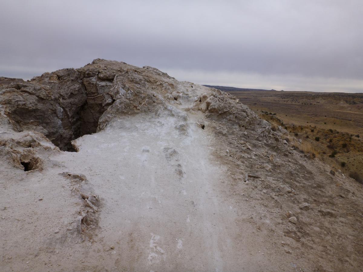 A rocky terrain with a wide, rugged path leading up to a summit, surrounded by dry, expansive landscape under a cloudy sky. The ground is light-colored and uneven, with some crevices and small holes present in the rock. White Ridge Bike Trails mountain bike trail.