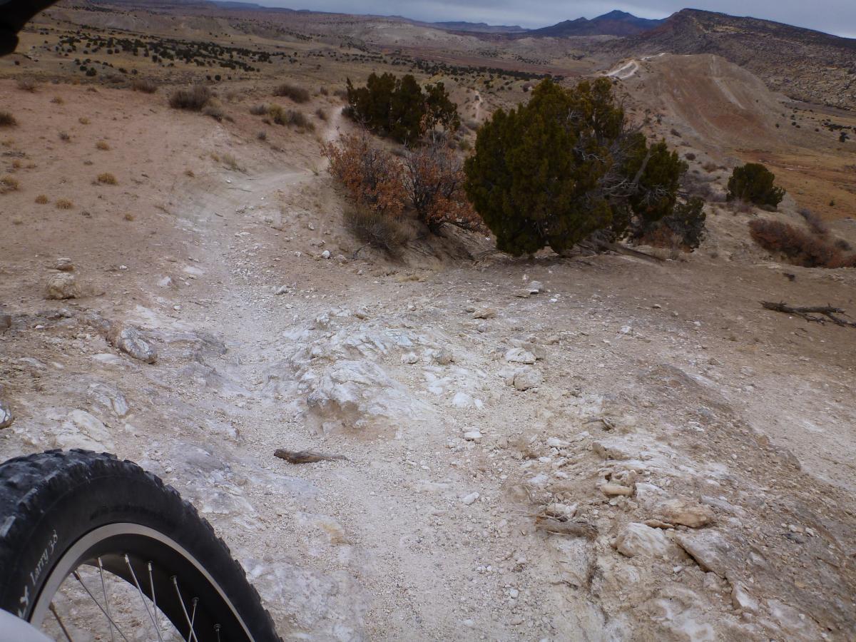 A mountain bike trail winding through a rugged, arid landscape with rocky terrain and sparse vegetation. In the foreground, a portion of a bike tire is visible, while the trail leads downhill into the distance, surrounded by hills and scattered trees. White Ridge Bike Trails mountain bike trail.