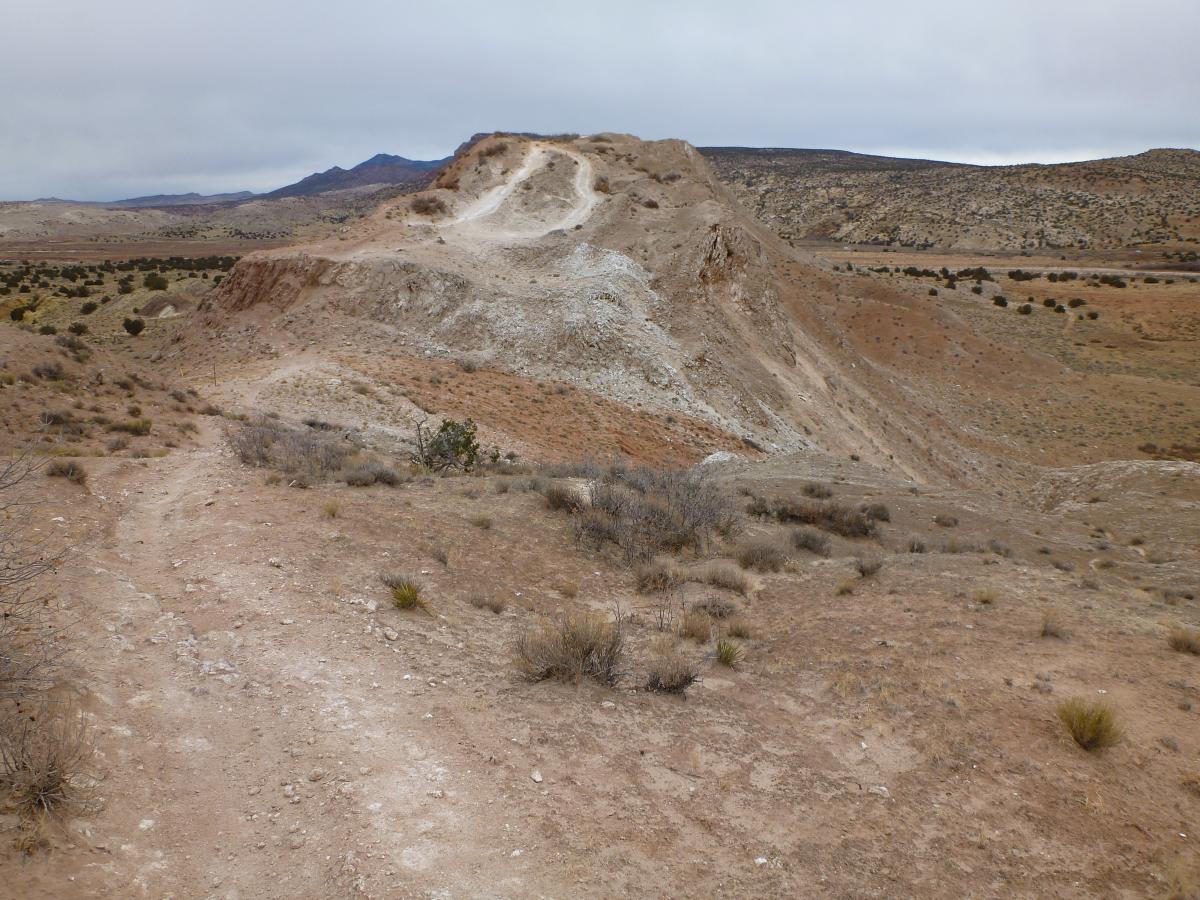 A rugged landscape featuring a dirt trail winding its way up a rocky hill surrounded by arid terrain. The hillside is marked by erosion, with a mix of brown and gray soil, and sparse vegetation dotting the landscape. In the background, rolling hills and mountains stretch into the distance under a cloudy sky. White Ridge Bike Trails mountain bike trail.