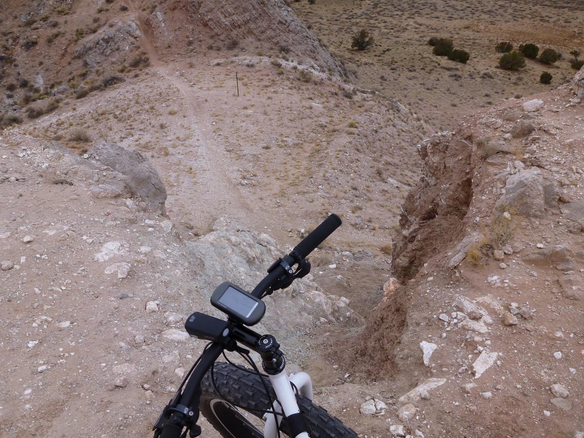 A view from the top of a rocky hillside, showing the handlebars of a mountain bike. The rugged terrain slopes down steeply in the background, with a dirt path visible leading away into the distance. The landscape is dry and sparsely vegetated, with scattered bushes and rocky outcroppings. White Ridge Bike Trails mountain bike trail.