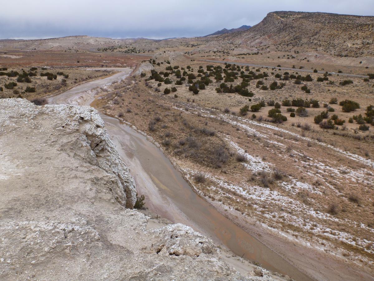 A panoramic view of a dry, rugged landscape featuring a winding riverbed and scattered bushes. The scene is set under a cloudy sky, with distant hills and mesas visible in the background. The ground is rocky and barren, showcasing earthy tones of brown and gray typical of arid environments. White Ridge Bike Trails mountain bike trail.