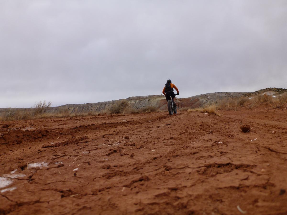 A cyclist riding a mountain bike along a rugged, dirt trail in a dry, rocky landscape under a cloudy sky. The terrain features cracked, reddish soil with sparse vegetation in the background. White Ridge Bike Trails mountain bike trail.