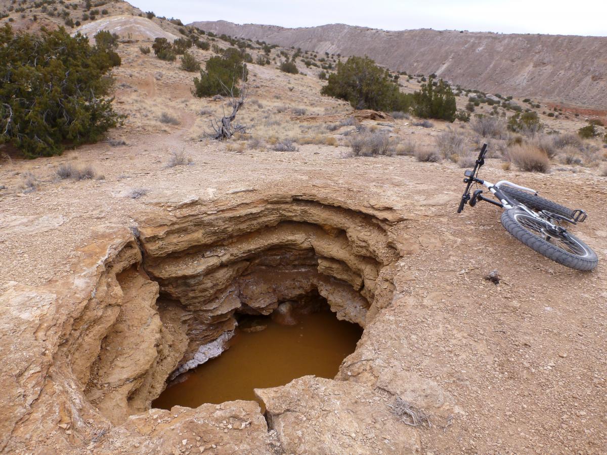 A dried, rocky landscape with a large, circular hole in the ground revealing brown water at the bottom. Nearby, a mountain bike rests on its side, partially obscured by the terrain. Sparse vegetation, including small bushes and trees, surrounds the area, with distant hills visible in the background under a cloudy sky. White Ridge Bike Trails mountain bike trail.