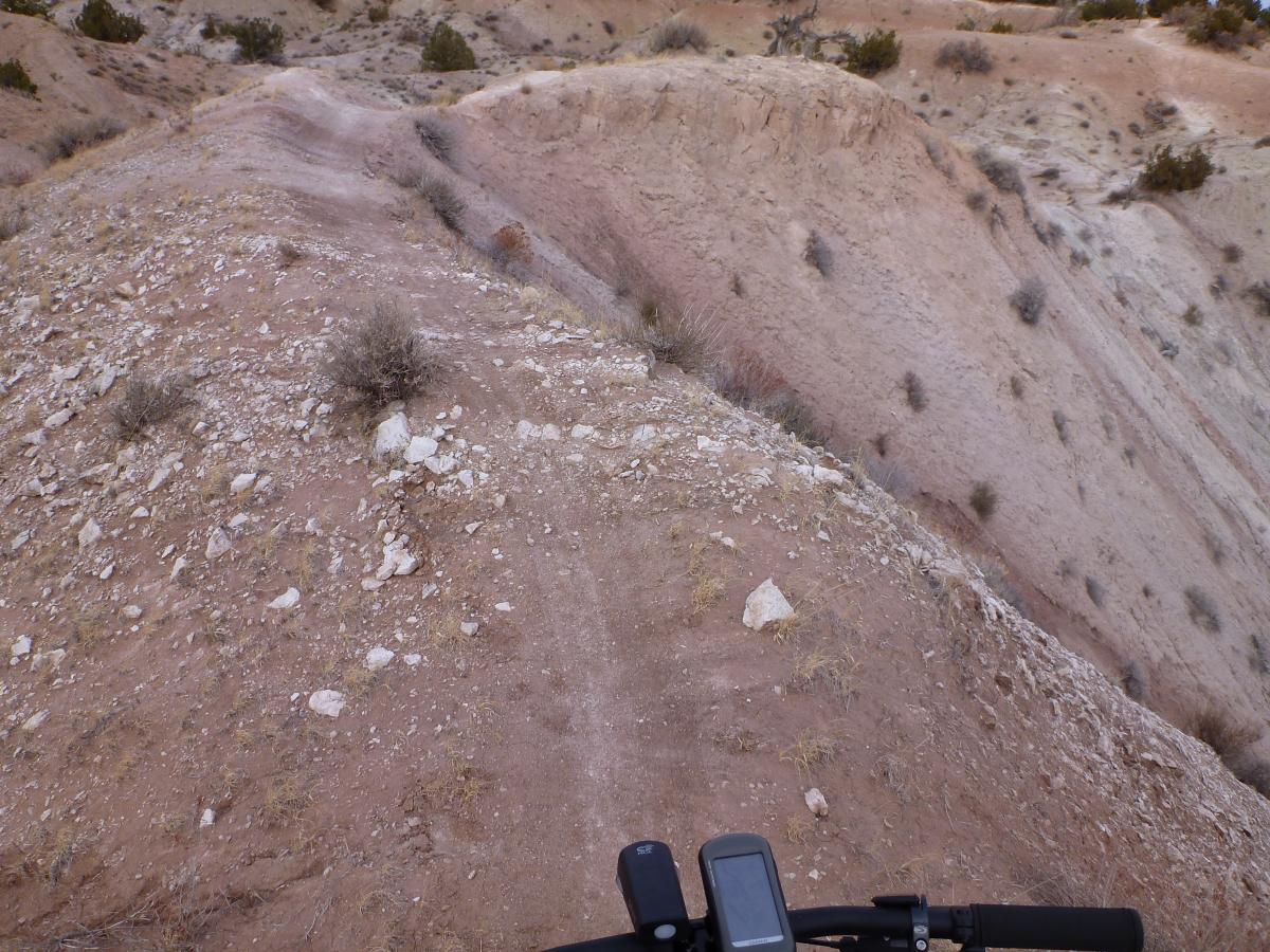 A rugged dirt trail winding along a steep, rocky hillside in a desert landscape, with sparse vegetation and varying shades of brown earth. The image captures the perspective from a bicycle's handlebars, hinting at an adventurous cycling route. White Ridge Bike Trails mountain bike trail.