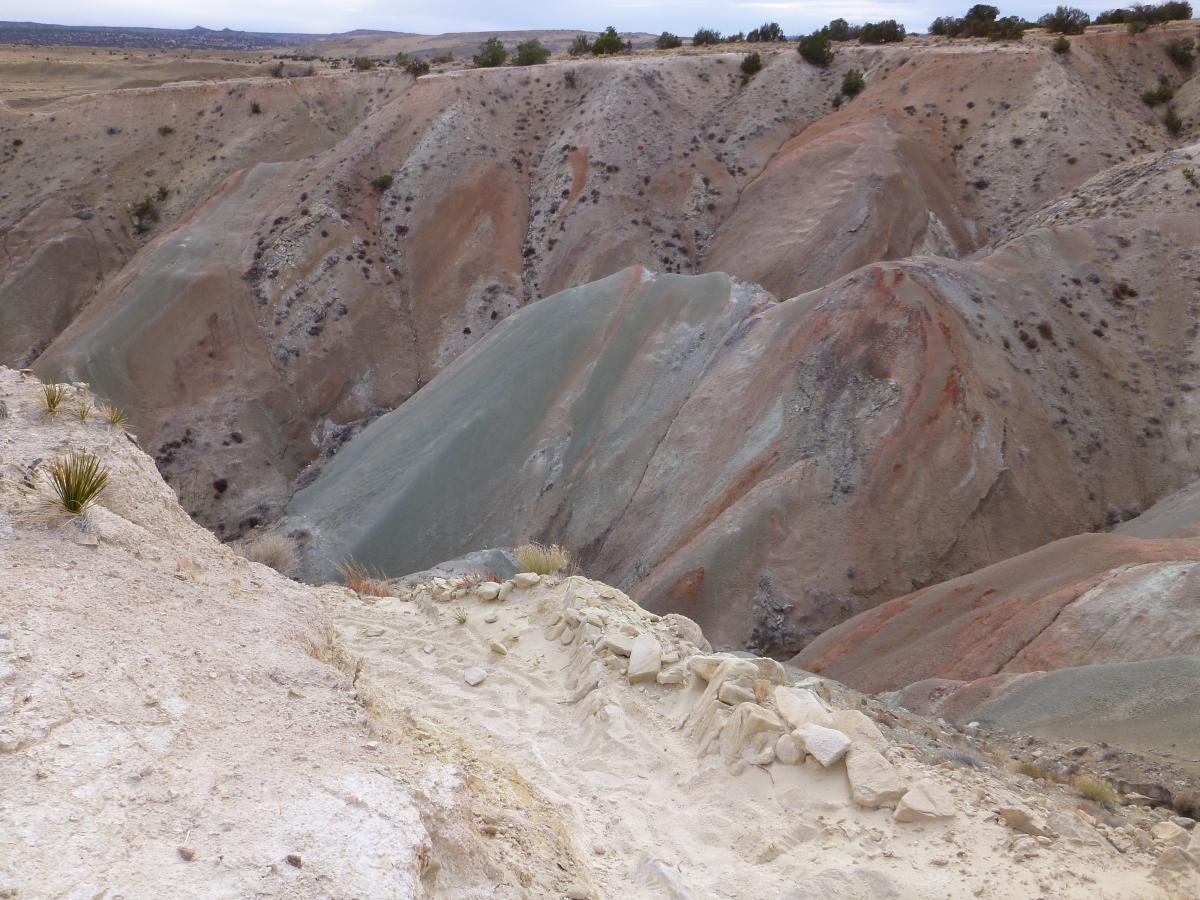 A scenic view of colorful, undulating hills and canyons, showcasing layers of reddish and greenish earth tones. The foreground features a rocky ledge with sparse vegetation, leading down into the rugged terrain below. A cloudy sky looms above, adding to the dramatic landscape. White Ridge Bike Trails mountain bike trail.