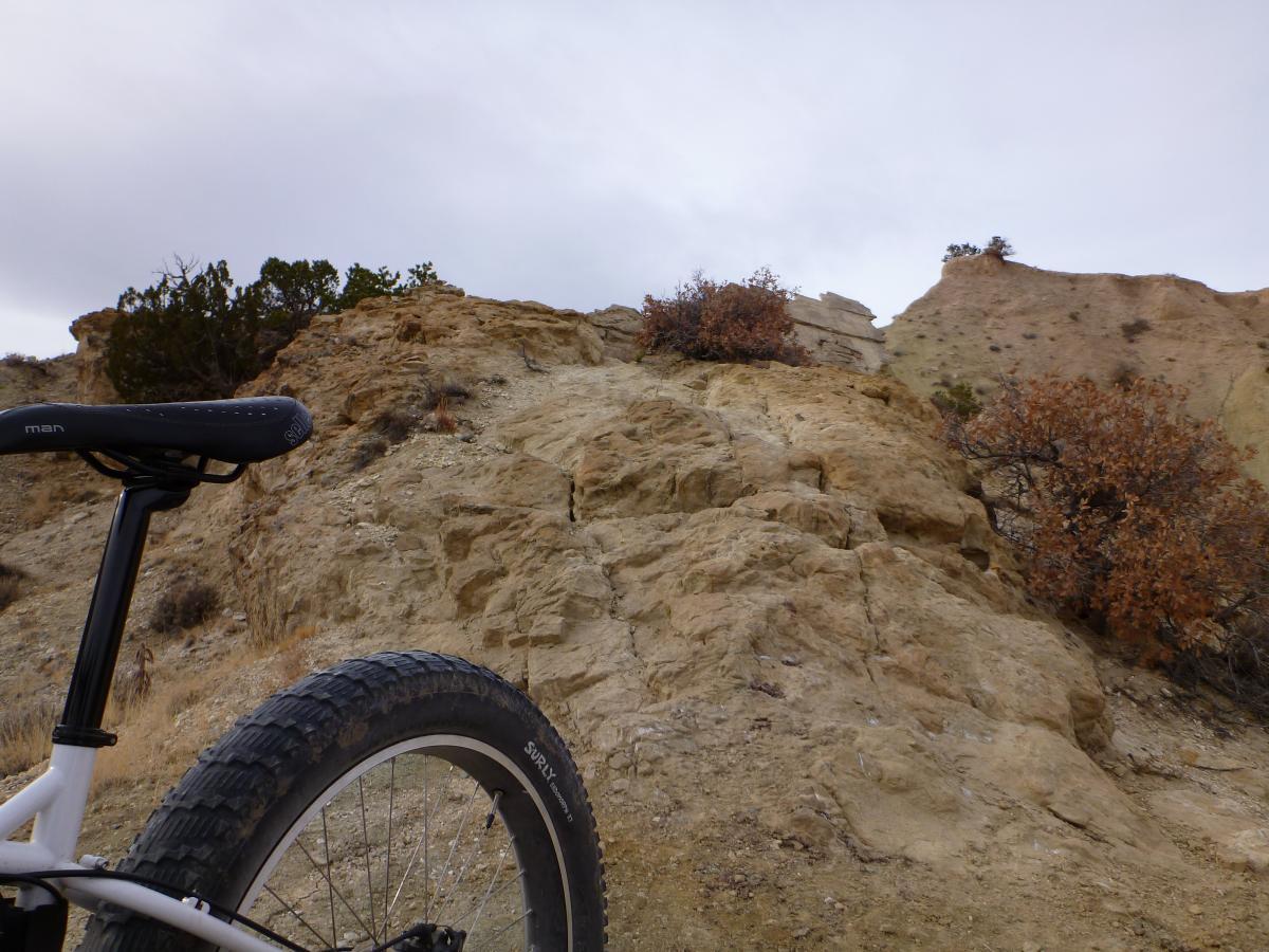 A rugged landscape featuring a close-up view of a mountain bike with a thick tire, positioned at the base of a rocky incline. The background showcases steep, eroded cliffs and sparse vegetation under a cloudy sky. White Ridge Bike Trails mountain bike trail.