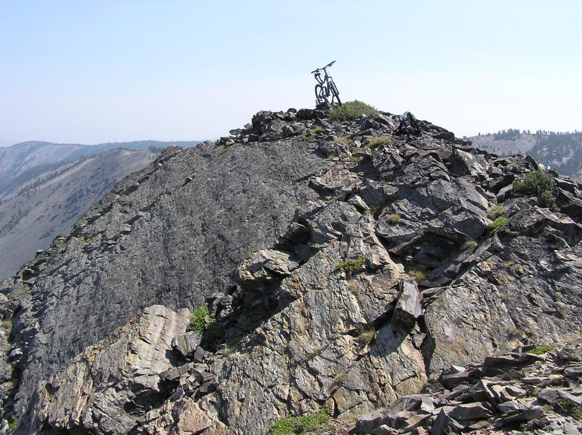 A mountain bike placed on top of a rocky, rugged peak with a panoramic view of distant mountains and a clear sky. The terrain is uneven with patches of greenery amidst the rocky surface. Eunice, Lake, Park, Goldstone, CDT Loop mountain bike trail.