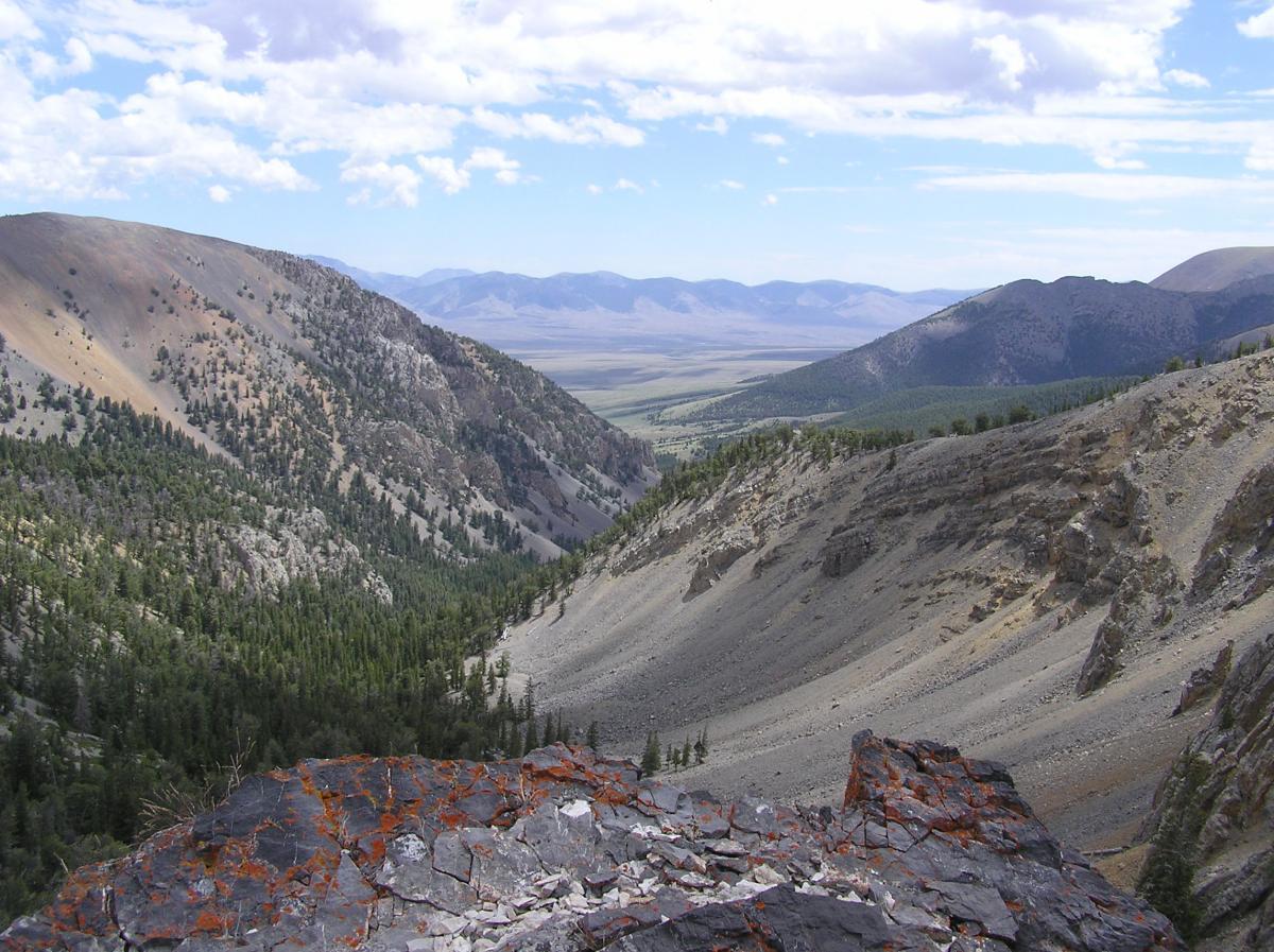 A panoramic view of a mountainous landscape, featuring rugged hills and valleys. The foreground shows a rocky outcrop with reddish lichen, while lush green forests cover the slopes. In the distance, expansive valleys and layered mountain ranges stretch under a clear blue sky dotted with white clouds. Mammouth Canyon Trail mountain bike trail.